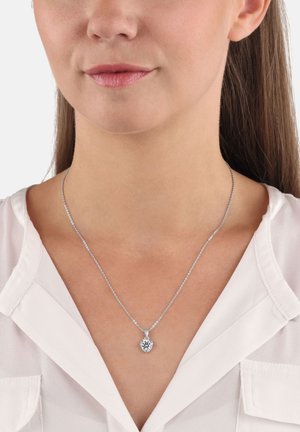 Collana d'argento con una delicata catena e un pendente a forma di cuore con un gemma trasparente, indossata sulla pelle nuda appena sopra una camicia bianca.
