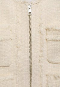 Cremefarvet jakke med et tekstureret stof, der har en central metallisk lynlås og to frontlommer med frynset kant.