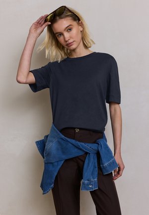 Jonge vrouw die een donker T-shirt draagt met een blauwe spijkerjassen om haar middel gestrikt, tilt een zonnebril van haar hoofd tegen een effen achtergrond.