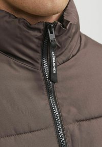 Giubbotto imbottito marrone con colletto alto, caratterizzato da una zip nera e un tirazip con marchio. Il tessuto ha una texture liscia e opaca.