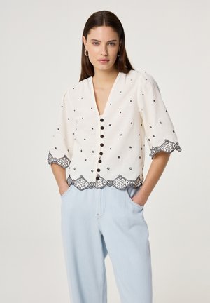 Frau trägt weiße, geknöpfte Bluse mit schwarzer Fischstickerei und wellenförmigen Kanten, kombiniert mit hellblauen, hoch taillierten Hosen, Hände in den Taschen.