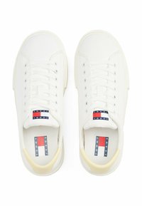 Baskets en toile blanches avec un bout rond et une semelle en caoutchouc. Elles possèdent des œillets en argent, des lacets plats blancs, et une étiquette de logo en rouge, blanc et bleu.