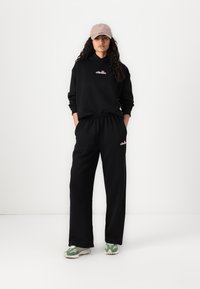 Čierna mikina s logom na hrudi, doplnená čiernymi teplákmi. K outfitu patrí béžová čiapka a zeleno-biele tenisky.