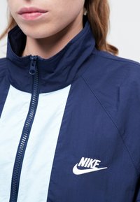 Mornarsko plava i svijetloplava Nike jakna s patentnim zatvaračem, visokim ovratnikom i izvezenim logotipom na prsima. Ima laganu, glatku teksturu.