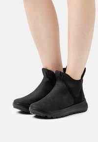 Bottines noires avec une texture lisse et mate, dotées de panneaux latéraux élastiques et de soufflets pour un enfilage facile. Semelle légère et design minimaliste.