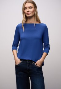 Blaues Langarmshirt mit Boot-Ausschnitt und weicher Textur, kombiniert mit dunklen Jeans, Hände in den Taschen. Einfaches und modernes Design.