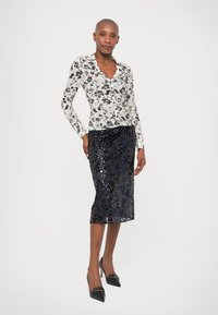 Woman standing with hand on hip wearing a white floral long-sleeve top, black sequined midi skirt, and black heels on white background.