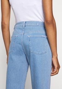 Vaaleansiniset farkut korkealla vyötäröllä, joissa on viisi taskua ja ompeleita korostavia yksityiskohtia. Kankaassa on sileä, hieman haalistunut rakenne.