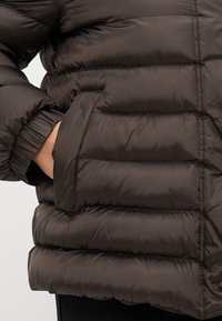 Rjava pufi jakna z žakardnim vzorcem, elastičnimi manšetami in stranskimi žepi. Material je sijoč in gladek, z lahkotnim občutkom.