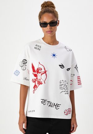Donna che indossa una maglietta bianca oversize con cupido rosso, occhi neri, sole blu, dadi e vari testi e simboli a tema amore.