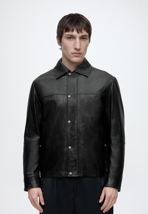 Άνδρας φοράει μαύρο δερμάτινο σακάκι με κουμπιά και γιακά, στέκεται μπροστά σε απλό ανοιχτό φόντο, κοιτώντας ευθεία με ουδέτερο ύφος.