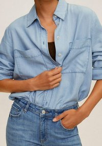 Camisa de mezclilla azul claro con cuello, dos bolsillos en el pecho y cierre de botones, combinada con jeans azul claro, usada de manera informal.