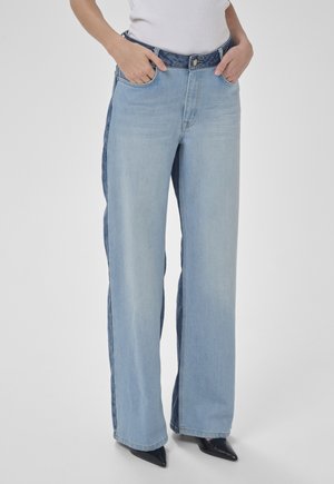 Osoba nosi svetlomodré džíny s širokými nohavicami s tmavomodrými bočnými panelmi, ruky v vreckách, v kombinácii s čiernymi špicatými topánkami a bielym tričkom.