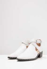 Botas blancas hasta el tobillo con punteras puntiagudas, acentos de tachuelas y una correa decorativa. Fabricadas en cuero suave con un tacón bajo y suela negra.