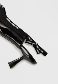 Čierna lodička na vysokom opätku z lakovanej kože so špicatou špičkou, s čiernym elastickým remienkom s nápisom "KARL LAGERFELD" a elegantným siluetou.