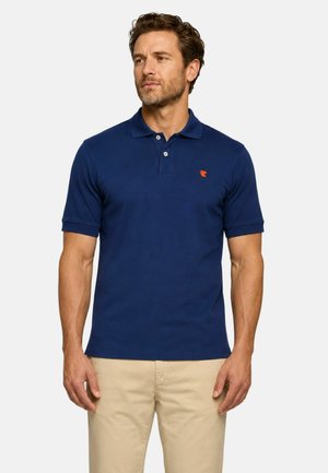 Uomo che indossa una polo blu navy a maniche corte con logo arancione e pantaloni beige, in piedi davanti a uno sfondo chiaro e uniforme.