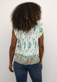 Femme aux cheveux bouclés portant un haut sans manches crème avec des motifs floraux teal et une bordure décorative brune, associé à un jean bleu.