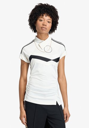 Mujer con cabello rizado que lleva una camiseta blanca de manga corta con patrones geométricos negros y pantalones negros, de pie frente a un fondo sencillo.