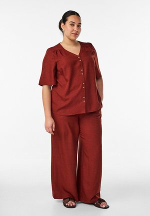 Donna in piedi, indossa un top color ruggine con scollo a V e bottoni, maniche al gomito e pantaloni larghi abbinati, sandali neri visibili.