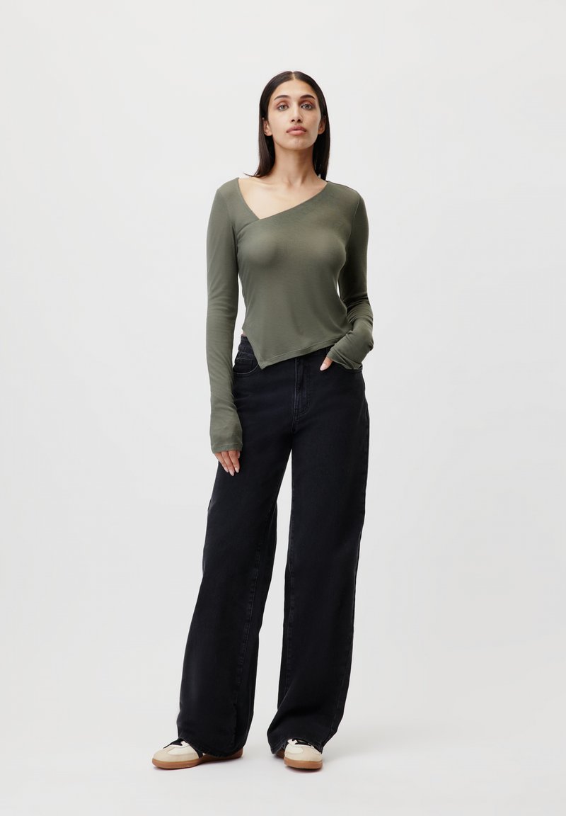 Top a maniche lunghe di colore verde oliva con scollatura asimmetrica, abbinato a jeans neri a vita alta e larghi e sneakers chiare.