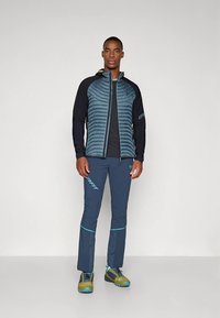 Heren outdoorjack met een donkerblauwe doorgestikte romp, zwarte mouwen en groene accenten. Gecombineerd met bijpassende broek en duurzaam schoeisel.