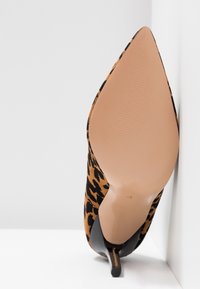 Chaussure à talon haut à bout pointu avec dessus imprimé léopard et semelle beige lisse, positionnée debout contre un mur blanc d'angle.
