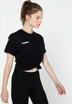 Schwarzes T-Shirt mit kurzen Ärmeln und geknotetem Vorderdesign, mit einem weißen "hummel"-Logo auf der linken Brust. Tragen mit schwarzen Leggings.