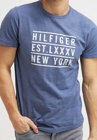 Hombre con una camiseta azul con el texto blanco "HILFIGER EST. LXXXV NEW YORK" de pie con una mano en el bolsillo.