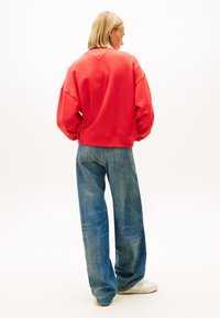 Červená oversized mikina s rebrovanými manžetami, v kombinácii s voľnými modrými džínsami a bielymi teniskami, zobrazená zozadu na jednoduchom pozadí.