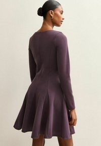 Mujer con el cabello en un moño, vistiendo un vestido oscuro morado de manga larga y acampanado, mirando de lado contra un fondo claro y liso.