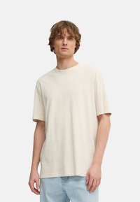 Camiseta de algodón color crema con cuello redondo, mangas cortas y un sutil logo en el pecho. Textura suave, corte relajado.