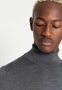 Grå turtleneck sweater lavet af ribbet strikket stof, med en høj krave og tætsiddende design. Teksturen er glat med subtile detaljer.