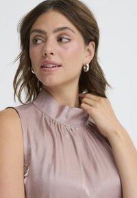 Blusa senza maniche di un delicato rosa con colletto alto, caratterizzata da un design plissettato; il modello tocca il colletto indossando orecchini a cerchio in argento.