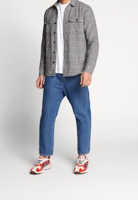 Man som bär en grå rutig skjorta med knappar, vit t-shirt, blå jeans och vita sneakers med röda och svarta detaljer.