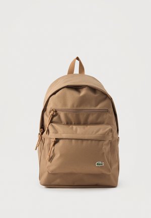 Sac à dos en tissu couleur fauve avec une poignée supérieure, un compartiment principal zippé, une poche avant zippée et un petit logo crocodile, présenté sur un fond uni.