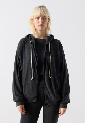 Heledate juustega inimene, kes kannab üle suurusega tumehallist luku ja valgete nööridega kapuutsi, seisab ühevärvilise heleda tausta ees.