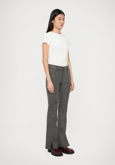 Γυναίκα που στέκεται με το πλάι, φοράει λευκή κοντομάνικη μπλούζα, γκρι ριγέ παντελόνια με ανοιχτές μύτες μπροστά και μπορντό παπούτσια.