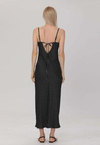 Robe maxi noire à pois blancs avec des bretelles fines, dos ouvert avec détail à nouer, et une silhouette fluide. Le tissu semble lisse et léger.