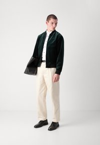 Chaqueta bomber de terciopelo verde oscuro, camisa blanca con botones negros, pantalones crema. Los accesorios incluyen un bolso negro y zapatos negros. Textura suave.