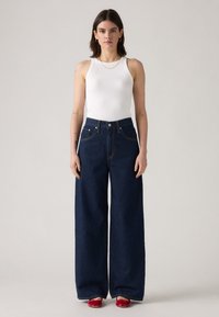 Temno modre kavbojke širokih hlačnic z visokim pasom, velikimi sprednjimi žepi in kontrastnim šivom, kombinirane s prilagojeno belo majico brez rokavov in rdečimi čevlji.