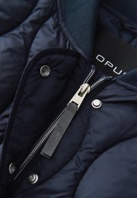 Giacca puffa navy con texture trapuntata, zipper metallico, bottoni a pressione e un gancio per appenderla. L'etichetta riporta "OPUS." Tessuto liscio e leggero.