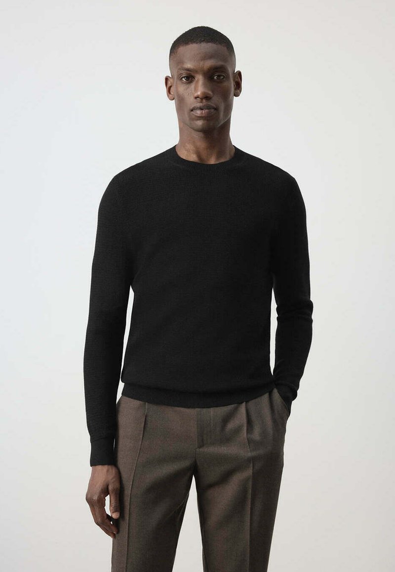 Homme portant un pull noir à manches longues et un pantalon plissé marron, debout avec une main dans la poche devant un fond uni.