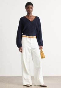 Navy gebreide trui met een V-hals, losse mouwen; witte hoge taille wijdpantalon. Gele riem en kleine gele handtas. Neutrale schoenen.