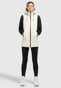 Helles beiges, gepolstertes Weste mit Kapuze, Reißverschluss und Seitentaschen, über einem schwarzen Langarmoberteil und schwarzen Leggings getragen, kombiniert mit weißen Turnschuhen.