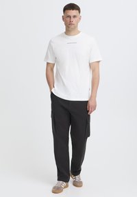 Hvid bomulds-T-shirt med tekstdetalje på brystet, kombineret med sorte cargobukser med sidelommer og en lige ben-design. Brune sneakers.