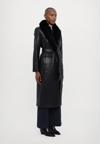 Μαύρο δερμάτινο παλτό με ψεύτικο γούνινο γιακά, ζώνη στη μέση και μακριά μανίκια. Φορεμένο πάνω από σκούρες φαρδιές παντελόνες και μαύρες μπότες με τακούνι.