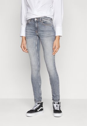 Personne portant un jean skinny gris clair, une chemise blanche et des baskets montantes noires avec des lacets et des semelles blanches, debout devant un fond blanc uni.