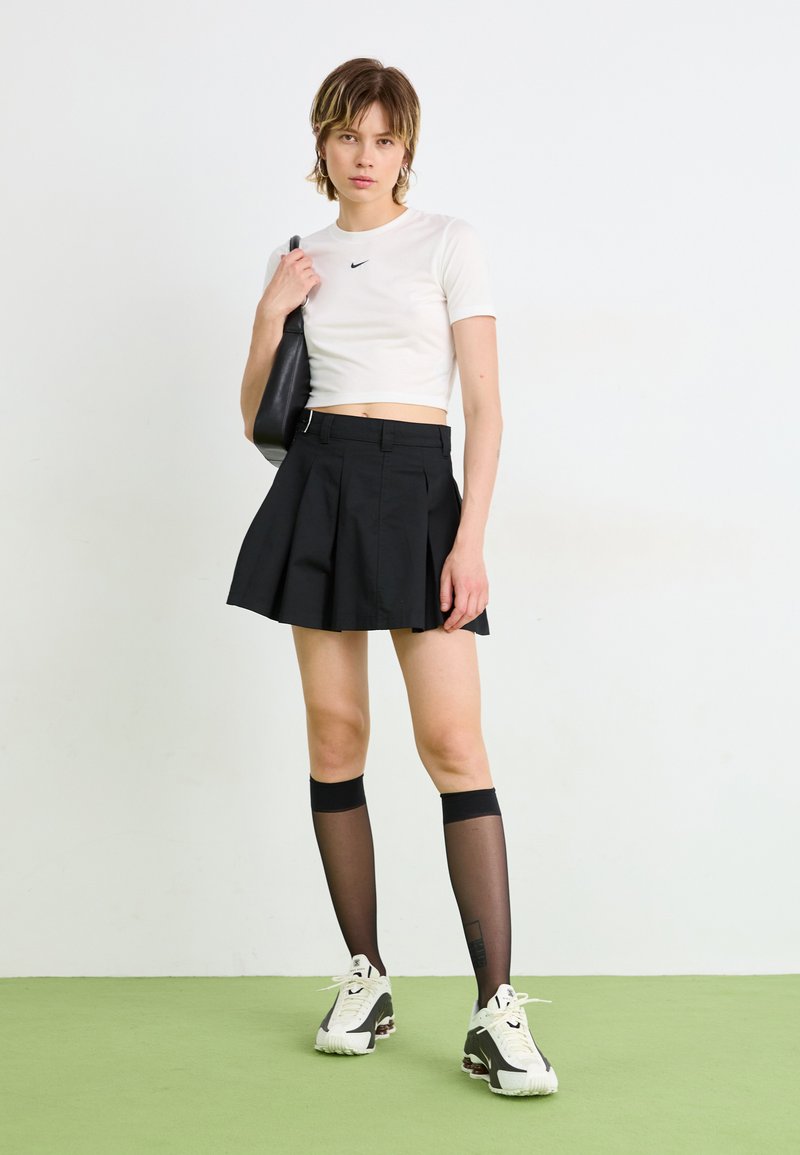 Weißes Nike-Crop-Top, schwarzer Faltenrock, schwarze knielange Socken und weiße Sportschuhe mit schwarzen Akzenten; vor einem hellen Hintergrund.