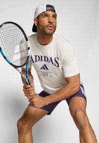 Adidas tennisantrekk med en kremfarget T-skjorte med lilla logo og lilla shorts. Holder en svart og blå tennisracket.
