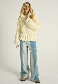 Žena s dlhými blond vlasmi v hrubom krémovom pletenom svetri na zips, dvojfarebných širokých džínsach a hnedých topánkach stojaca na jednoduchom pozadí.